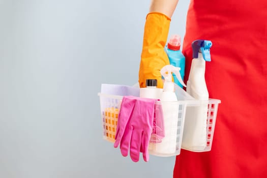 A woman in rubber gloves and red apron holding basket with sponge and cleaning products on blue background