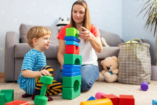 Cute little kid and child development specialist attractive young woman playing together with colorful blocks, sitting on the floor.