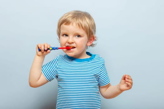 Little blonde boy brushing teeth with toothbrush on blue background. Dental hygiene.
