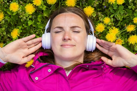 Beautiful young woman in headphones listens to music lying on the grass with dandelions on a spring day