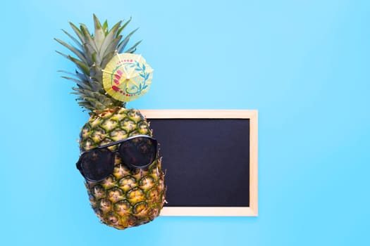 Pineapple in sunglasses with empty copy space blackboard for your text on blue background. Summer vacation party time concept.