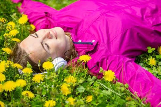 Beautiful young woman in headphones listens to music lying on the grass with dandelions on a spring day