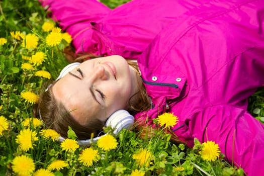 Beautiful young woman in headphones listens to music lying on the grass with dandelions on a spring day