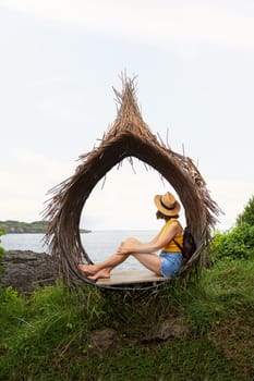 Young woman wearing hat relaxing sitting in straw nest looking at ocean view. Female contemplating sea landscape in Bali. Vertical image. Travel, vacation concept.