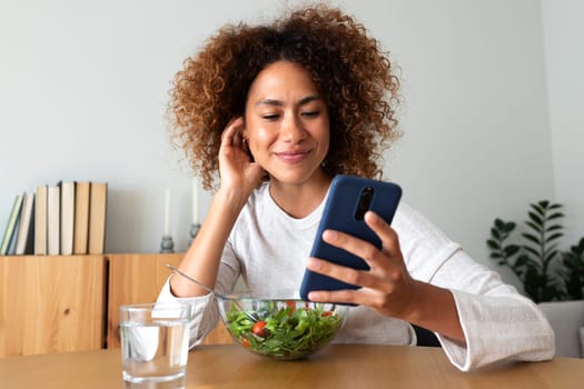 Young multiracial woman eating healthy salad while using phone at home. Healthy lifestyle concept.
