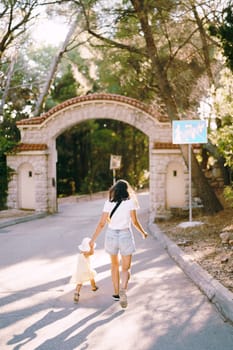 Mother with a little daughter run along the park road to the gate holding hands. Back view. High quality photo