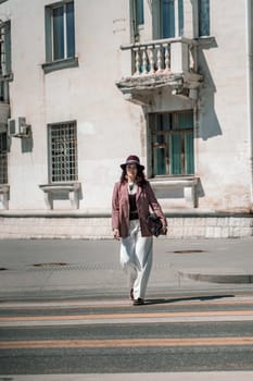 Woman city road crossing. Stylish woman in a hat crosses the road at a pedestrian crossing in the city. Dressed in white trousers and a jacket with a bag in her hands
