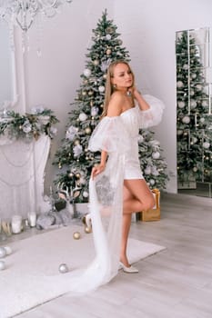 The blonde in the Christmas room. A beautiful blonde woman in a shiny light short dress with a train stands in a beautiful bright room decorated with a festive interior with a Christmas tree