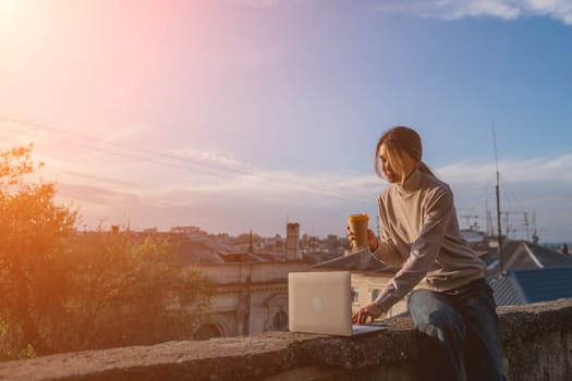 Woman freelancer uses laptop on cement wall outdoors against the sky and the roof of the city. The woman to be focused on her work or enjoying some leisure time while using her laptop