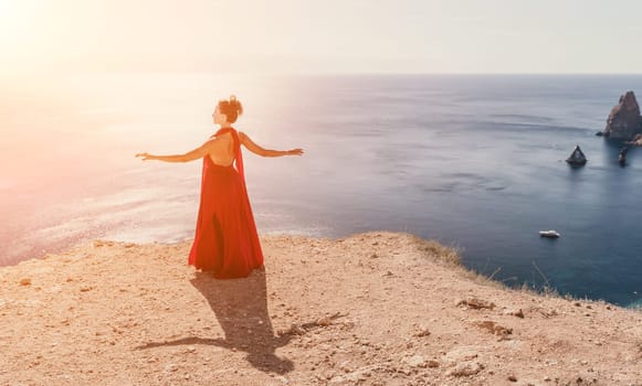 Side view a Young beautiful sensual woman in a red long dress posing on a rock high above the sea during sunrise. Girl on the nature on blue sky background. Fashion photo.