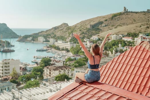 Woman sits on rooftop with outstretched arms, enjoys town view and sea mountains. Peaceful rooftop relaxation. Below her, there is a town with several boats visible in the water.