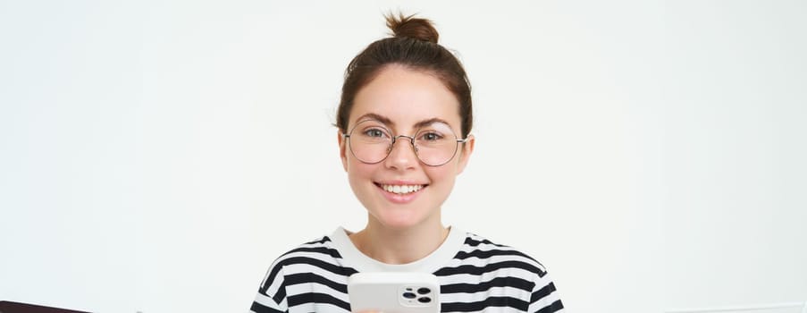 Portrait of beautiful young woman in eyewear, holding smartphone, using mobile phone, online shopping in application, standing over white background.