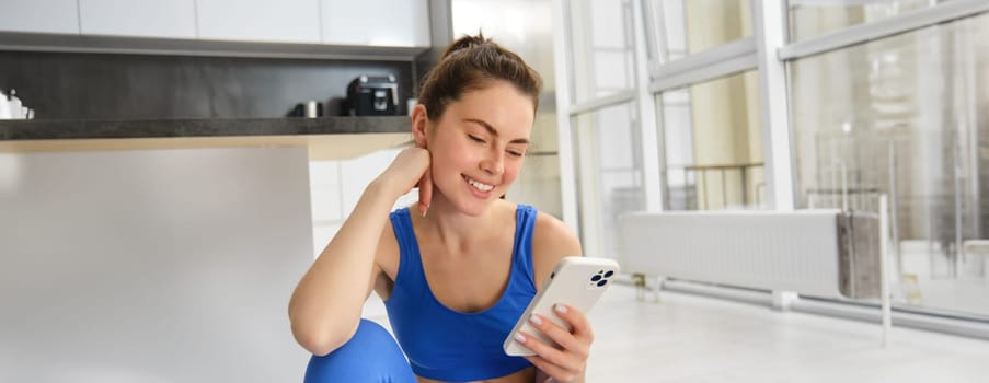 Image of young fit woman wearing sportbra and blue leggings, using smartphone app, does workout from home, sits on floor and follows online training video.