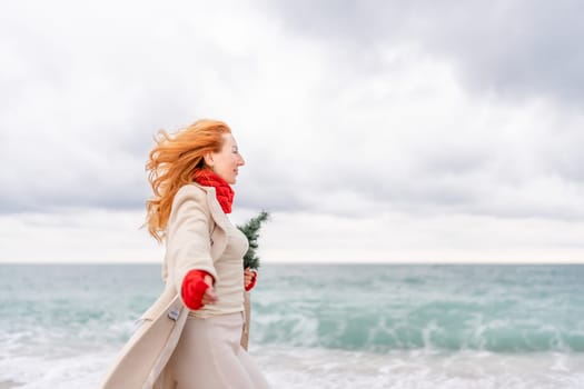 Redhead woman Christmas tree sea. Christmas portrait of a happy redhead woman walking along the beach and holding a Christmas tree in her hands. Dressed in a light coat, white suit and red mittens