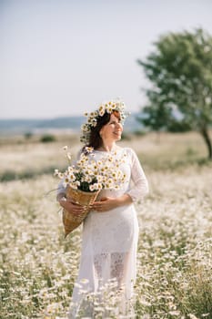 Happy woman in a field of daisies with a wreath of wildflowers on her head. woman in a white dress in a field of white flowers. Charming woman with a bouquet of daisies, tender summer photo.