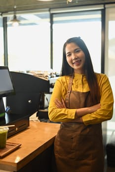 Portrait of small coffee shop owner standing with arms crossed behind counter with cash register.