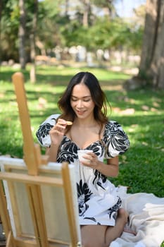 Happy young woman painting on easel sitting on the grass during a picnic in the park.