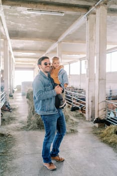 Little smiling girl sits in the arms of her father standing near the corral with goats. High quality photo
