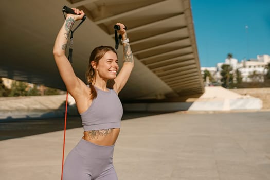 Young smiling woman makes fitness workouts with resistance band on morning city background