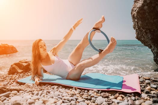 Young woman with black hair, fitness instructor in pink sports leggings and tops, doing pilates on yoga mat with magic pilates ring by the sea on the beach. Female fitness daily yoga concept