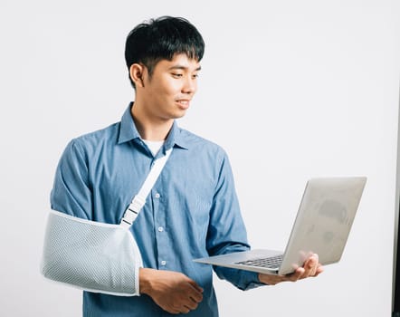 Injured Asian businessman, with a broken arm in a splint, remains dedicated to work on a laptop. Studio shot isolated on white, symbolizing resilience and recovery.