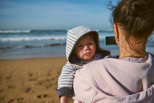Adorable baby boy in arms of his mother taking photo of sea on smartphone, standing on sandy beach back to camera and photographing beautiful waves splashing on the Atlantic ocean. People and nature
