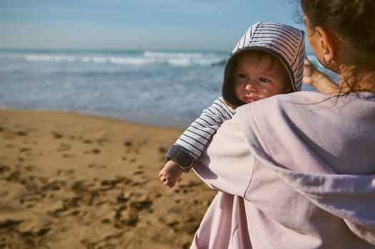 Authentic portrait of a Caucasian cute baby boy, in his mother's arms. Young mom carrying her lovely child while walking together on the beach. People and nature. Maternity leave. Babyhood and infancy