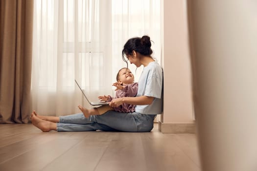 beautiful casual woman sitting on the floor and working on laptop with her little child girl at home