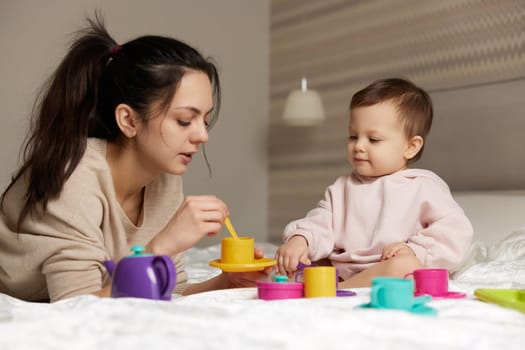 Attractive mother and little child daughter playing tea party and spending time together in bedroom, family having fun