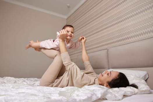 Happy Caucasian mother lifting in air cute little child girl and playing together in bedroom