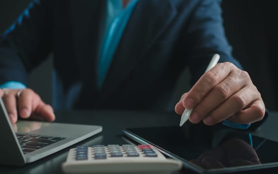 Businessman in black suit working on laptop computer and tablet and calculator, Hand Hands touch on tablet at office with dark background, Online working, Close up, Copy space.