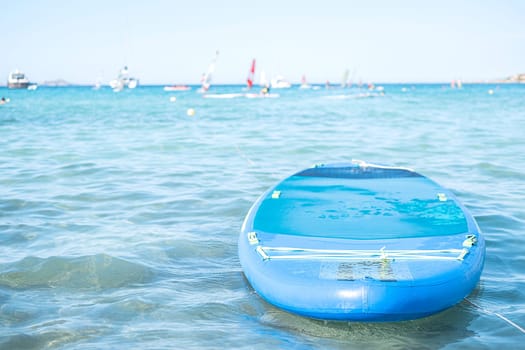 Lonely blue sup board on the sea on floating sailboats background on sunny day.