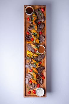 A long wooden platter filled with an assortment of meats and vegetables, including steak, sausage, chicken, potatoes, carrots, and peppers.