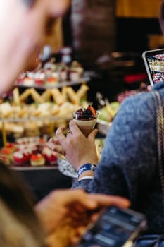 A person holds a dessert cup with chocolate mousse and berries. Blurred background with desserts and people in the distance.