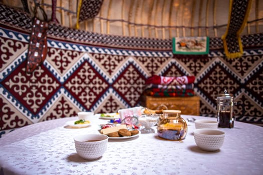 Inside a yurt, a table is set with cups, plates, and food. Colorful tapestries and carpets adorn the space.