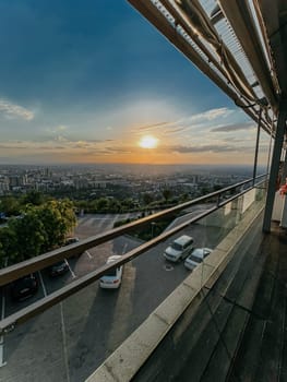 A beautiful sunset can be seen from a rooftop terrace with a glass railing. In the background, there is a parking lot and a city.