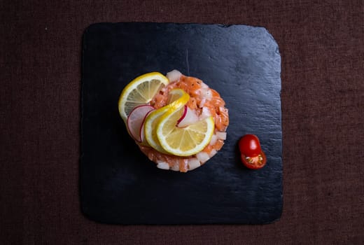 Savor a delightful salmon tartare with lemon, radish, and cherry tomato on a stylish slate plate for a burst of flavors and textures.