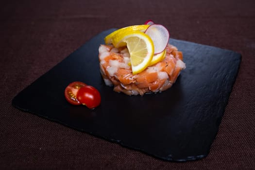 Savor a delightful salmon tartare with lemon, radish, and cherry tomato on a stylish slate plate for a burst of flavors and textures.