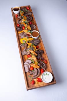 A long wooden platter filled with an assortment of meats and vegetables, including steak, sausage, chicken, potatoes, carrots, and peppers.