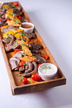 A long wooden platter filled with an assortment of meats and vegetables, including steak, sausage, chicken, potatoes, carrots, and peppers.