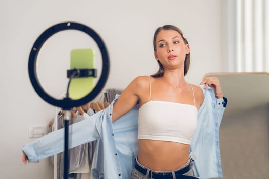 Young woman making fashion video content for social media. Blogger smiles to camera using selfie stick and light ring while making persuasive online clothing sell to audience or followers. Unveiling