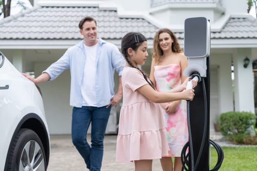 Happy little young girl learn about eco-friendly and energy sustainability as she help her family recharge electric vehicle from home EV charging station. EV car and modern family concept. Synchronos