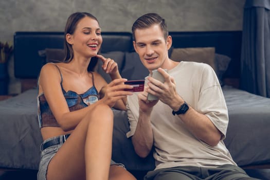 Young couple sit in the home bedroom using online payment app and digital wallet on smartphone to pay with credit card. E commerce shopping and modern purchasing via mobile internet. Unveiling