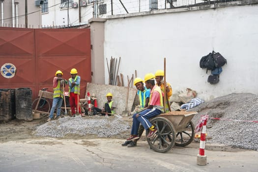 Antananarivo, Madagascar - April 24, 2019: Group of unknown Malagasy worker men in helmets and hi visibility jackets, resting at worksite, sitting and leaning to wheelbarrow, backpacks hanging on wall