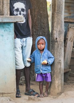 Ranohira, Madagascar - April 29, 2019: Unknown small Malagasy boy kid in blue denim jacked and shorts standing next to his father, holding bread in both hands. 