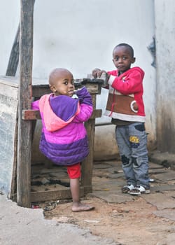 Ranohira, Madagascar - April 29, 2019: Two unknown Malagasy kids standing by empty wooden market stall on the street, looking into camera, one chewing piece of wood. 