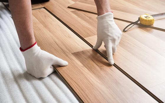 Installing laminated floor, detail on man hands in gloves fitting wooden tile, over white foam base layer, tape measure in background