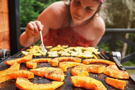 Butternut squash pieces grilled on electric grill, focus on bright orange vegetables seasoned with spice, blurred young woman in background