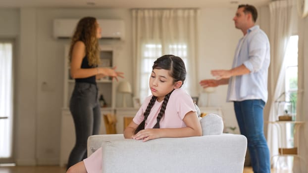 Annoyed and unhappy young girl sitting on sofa trapped in middle of tension by her parent argument in living room. Unhealthy domestic lifestyle and traumatic childhood develop to depression Synchronos