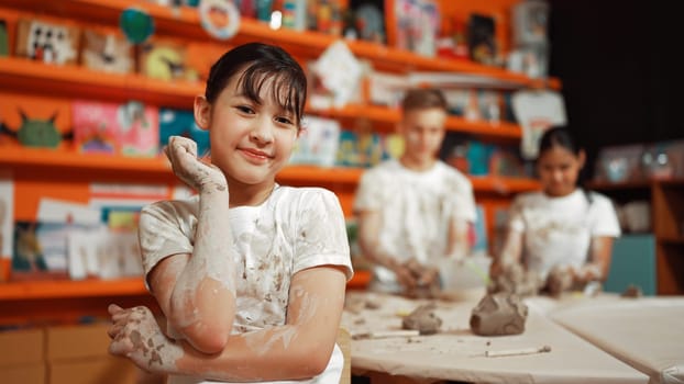 Asian highschool girl look at camera and dirty hand while diverse children modeling clay behind. Happy cute student wearing shirt while looking at camera at workshop. Blurring background. Edification.